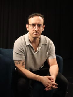 Headshot of Evan, a white man with short brown hair. He wears a taupe short-sleeved polo and a serious expression.