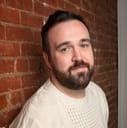 Headshot of Frank, a white person with short brown hair and beard, wearing a white sweater and leaning on a brick wall.
