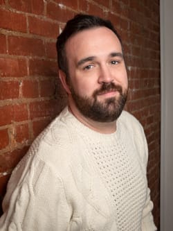 Headshot of Frank, a white person with short brown hair and beard, wearing a white sweater and leaning on a brick wall.