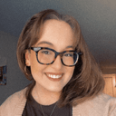 Isabel is a white woman in her 20s with light brown hair and glasses. She smiles brightly, wearing a peach cardigan.