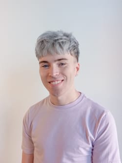 Headshot of Jake. He's a young white man with gray hair, smiling and wearing a light-colored tee against a white background.