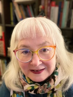 K.A. McFadden is a white person with blonde hair, bangs, & glasses with rainbow-hued frames. They wear a patterned scarf.