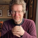 Photo of Kile Smith. He is a white man with graying beard and hair, smiling at a restaurant table, wearing a maroon shirt.