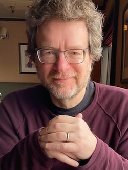 Photo of Kile Smith. He is a white man with graying beard and hair, smiling at a restaurant table, wearing a maroon shirt.