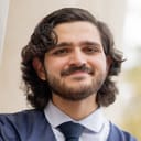 A young man with light skin and  wavy brown hair and a short beard and mustache. He is smiling and wears a suit and tie.