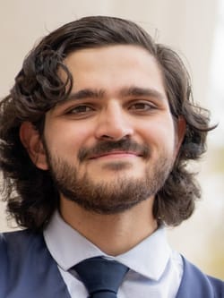 A young man with light skin and wavy brown hair and a short beard and mustache. He is smiling and wears a suit and tie.