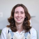 Headshot of Maggie, a smiling white woman wearing an argyle vest. She has shoulder-length wavy brown hair.