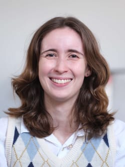 Headshot of Maggie, a smiling white woman wearing an argyle vest. She has shoulder-length wavy brown hair.