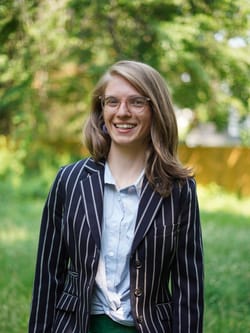 Sturges is a white woman with wavy blonde and glasses. She smiles and wears a pin-striped black blazer.