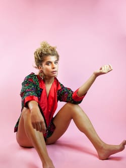 Sarah, a white woman with curly hair, in a silk jacket sans pants, sits nonchalantly on the floor on pink backdrop.