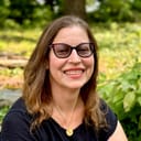 Gabrielle is a white woman in her 50s with brown hair and glasses, smiling brightly outdoors.