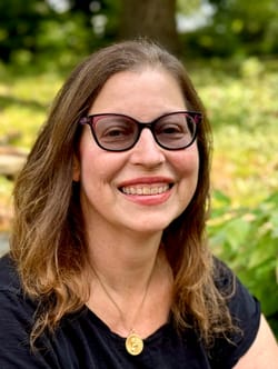 Gabrielle is a white woman in her 50s with brown hair and glasses, smiling brightly outdoors.