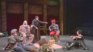 6 ensemble members in a motley range of costumes appear to be having a picnic on the black floor of the stage.