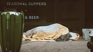 Photo of a person completely wrapped in a peach & blue colored blanket, lying on a city sidewalk next to a postal service bin