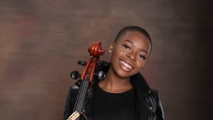 Ali, a Black woman, in all black with a black leather jacket, poses for a portrait next to a cello