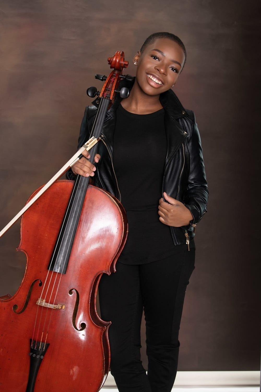 Ali, a Black woman, in all black with a black leather jacket, poses for a portrait next to a cello