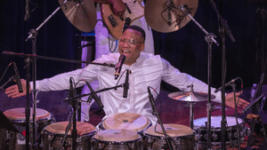 Martinez, an Afro-Cuban man, sits before congas, cymbals, and microphones, with his arms out, his mouth wide as he's sings.