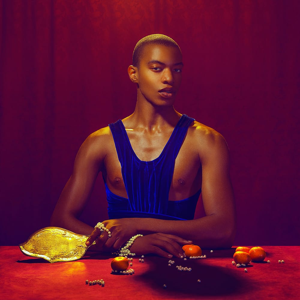 Portrait of a Black man in a blue strap top; jewelry beads and fruit on table in front of him. A red curtain behind him