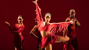 Dancers pose, dressed mostly in red with black and red undertones. One dancer is centered, three others are behind her.