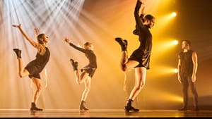 On a stage glowing with smoky white and yellow spotlights, 3 dancers in black boots throw their arms and right leg back.