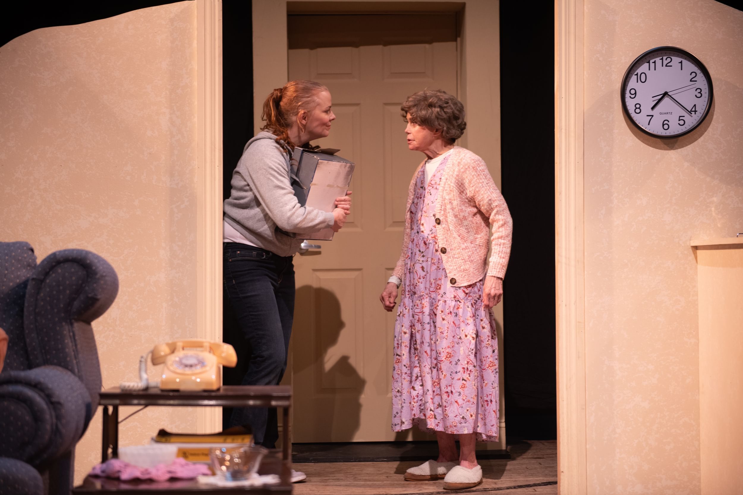 A scene from the play. Jessie holds a box in what looks like a bedroom door, facing her mom, who wears a floral house dress