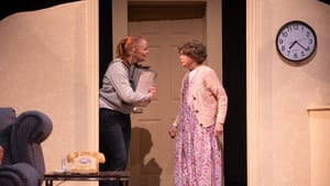 A scene from the play. Jessie holds a box in what looks like a bedroom door, facing her mom, who wears a floral house dress