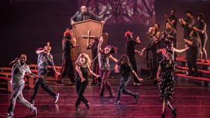 A scene from Lift. 6 people dance in unison in front of a wood pulpit with a cross. A gospel choir stands on risers at right