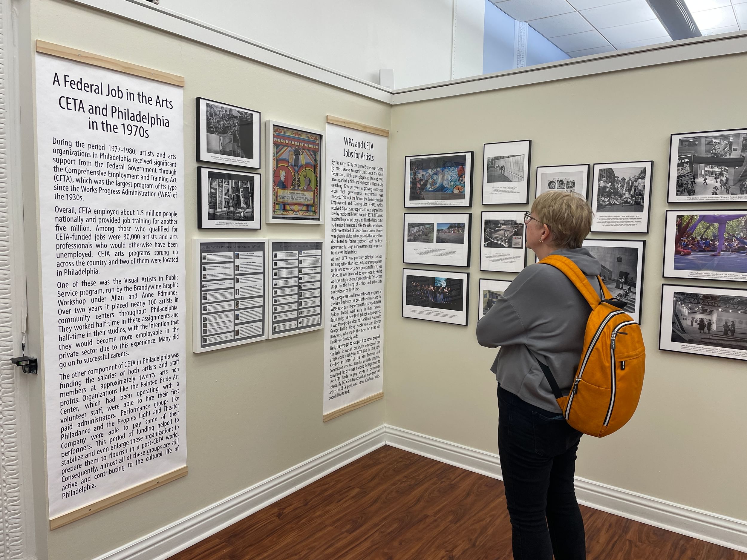 A woman with short hair and an orange backpack looks at text and photos in a gallery corner.