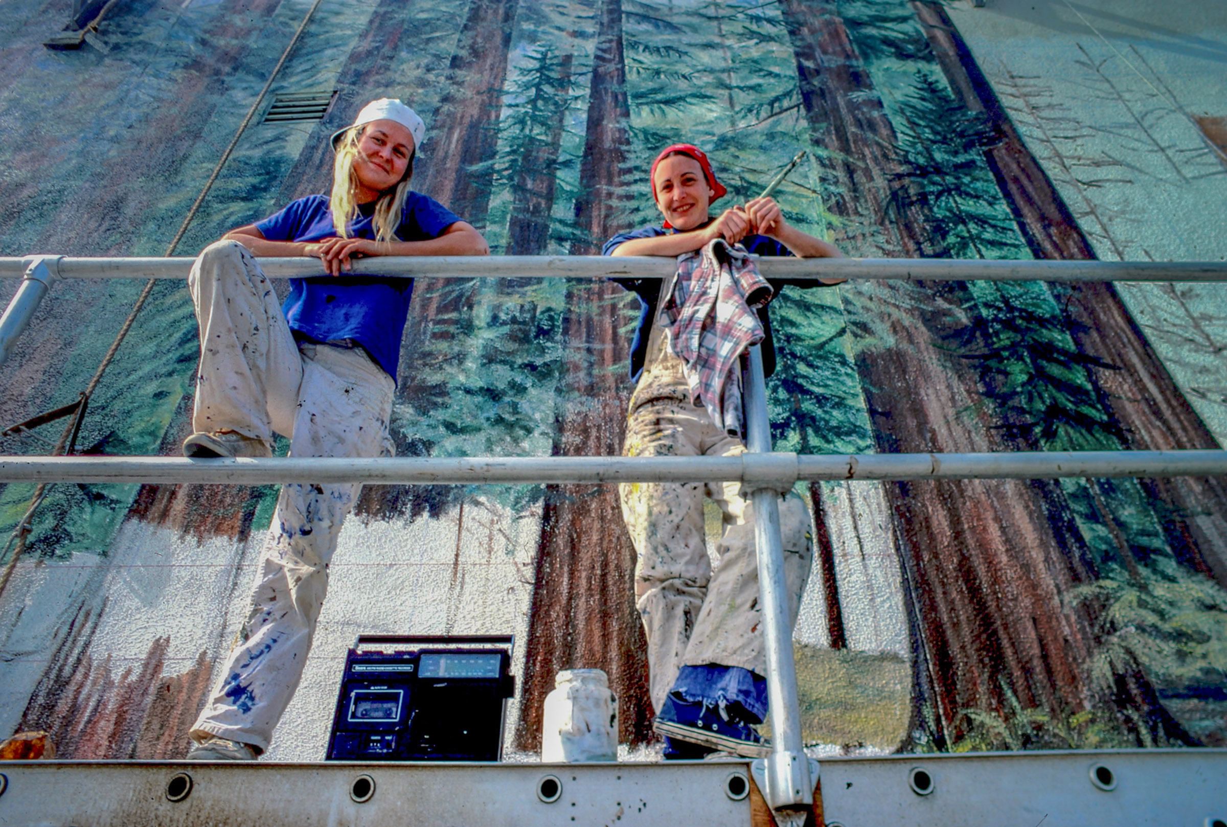 Two young white women in paint-spattered clothes pose happily against a railing in front of a mural of tall trees.