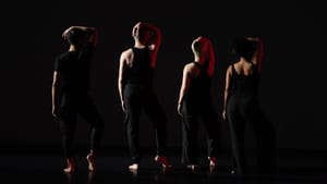 The four dancers, all in black, stand in a line facing away on a black stage, right arms bent upward to rest on their heads.