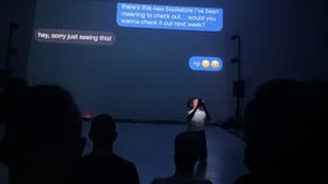 One actor kneels on a dark stage holding a smartphone. Their text convo projects behind, including “sorry just seeing this”