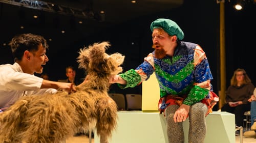 Elliot puppets a goofy, realistic life-sized large shaggy brown dog that Carpenter pets, wearing clashing colorful patterns