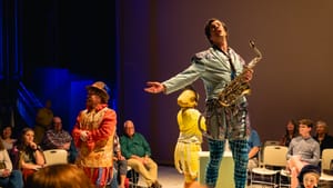 The two actors, in outlandish colorful Elizabethan-inspired clothes, perform surrounded by audience. Corey holds a saxophone