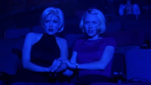 Two women with identical blonde bobcuts sit in a theater, slightly spooked, holding hands. The room is tinted an intense blue