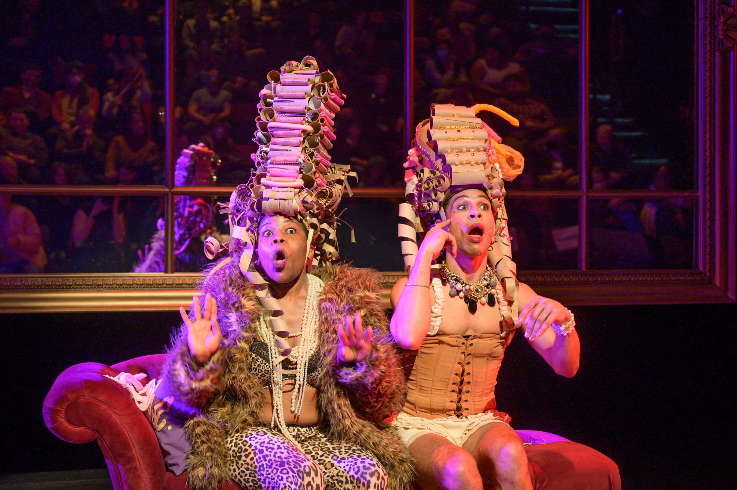 Kidwell and Kazen-Maddox sit on a couch looking shocked and wearing towering cardboard wigs like 18th-century French royalty