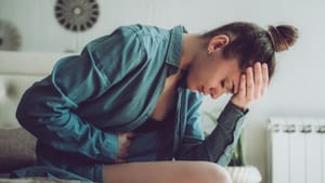 In profile, a young woman sits on the edge of a bed and leans over, clutching her head and stomach in pain.